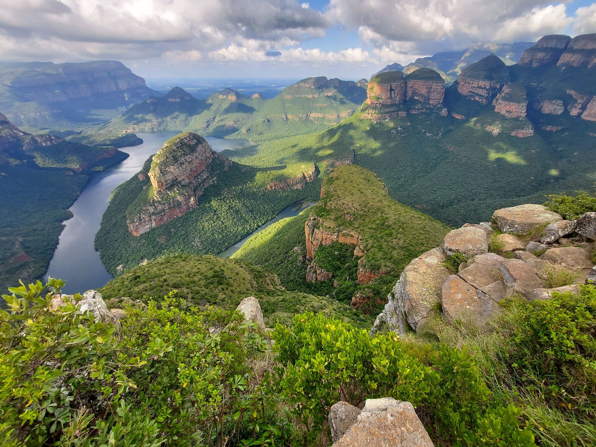 A deep river canyon seen from its rim, a winding river far below and flat-topped rock buttes along the far wall, all under dense green bush
