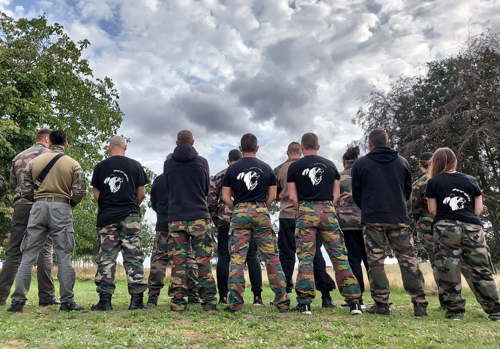 About fifteen people in camouflage and black T-shirts standing in a line with their backs to the camera, under a heavy grey sky